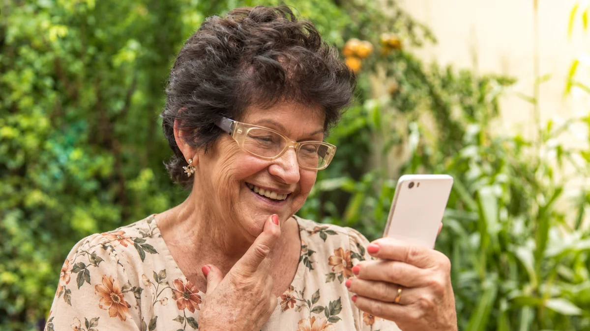 Senhora de óculos sorrindo enquanto olha o celular no quintal de casa