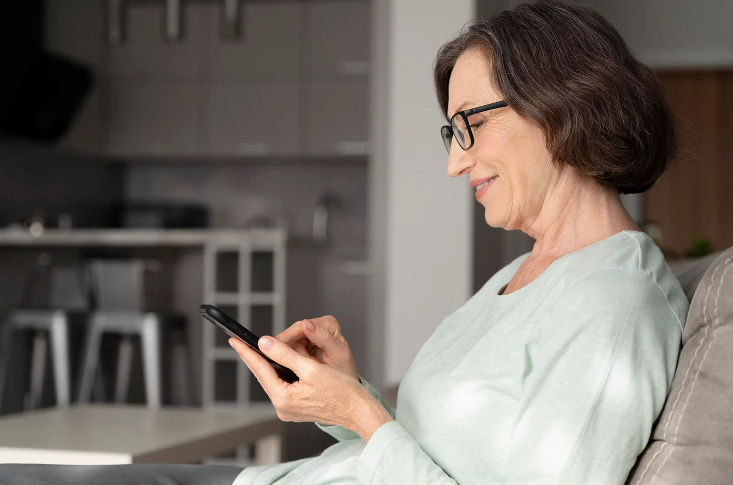 Senhora de óculos sorrindo enquanto olha o celular sentada no sofá