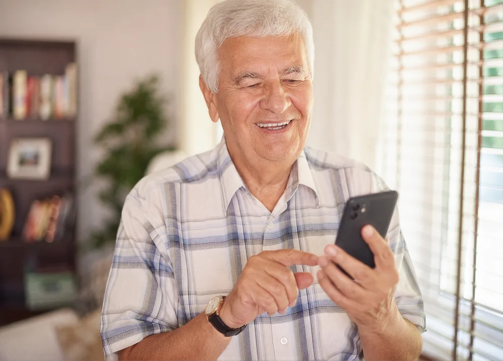Senhor de cabelos brancos sorrindo enquanto mexe no celular em casa